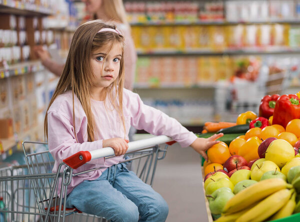 Cute surprised girl taking a fruit in the produce section at the supermarket, she is sitting on the shopping cart, healthy food concept