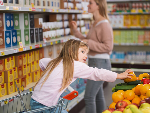 Smart girl sitting on the shopping cart and taking a fresh healthy fruit at the supermarket, her mother is standing in the background