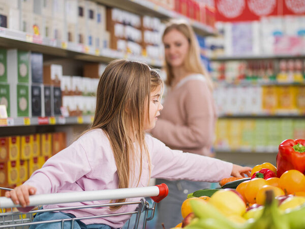 Smart girl sitting on the shopping cart and taking fresh healthy vegetables at the supermarket, her mother is standing in the background