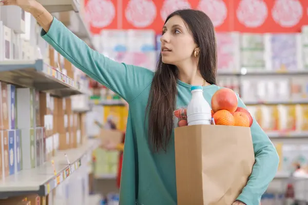 食料品店の棚から製品を取る若い魅力的な女性は 彼女は完全なショッピングバッグを保持しています — ストック写真