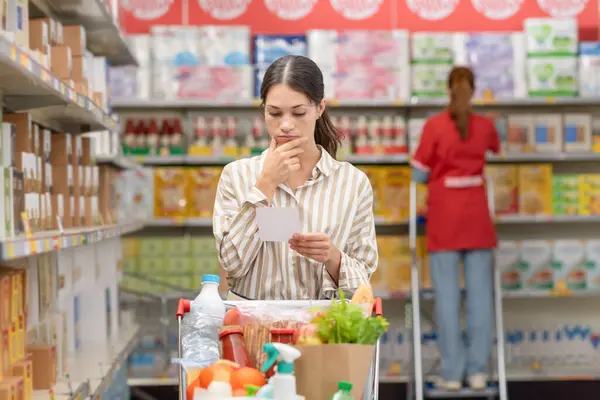 Mladá Zamyšlená Žena Plným Nákupním Vozíkem Supermarketu Kontroluje Svůj Nákupní — Stock fotografie