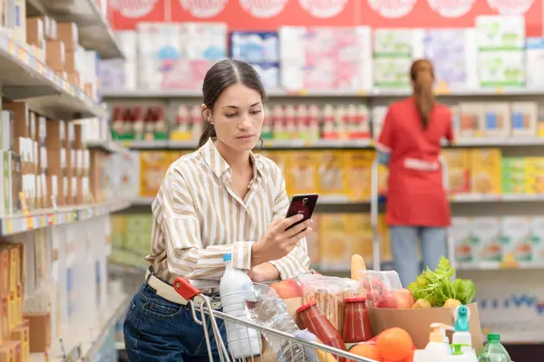 Žena Opírající Plné Nákupní Vozík Pomocí Svého Smartphonu Obchodě Potravinami — Stock fotografie