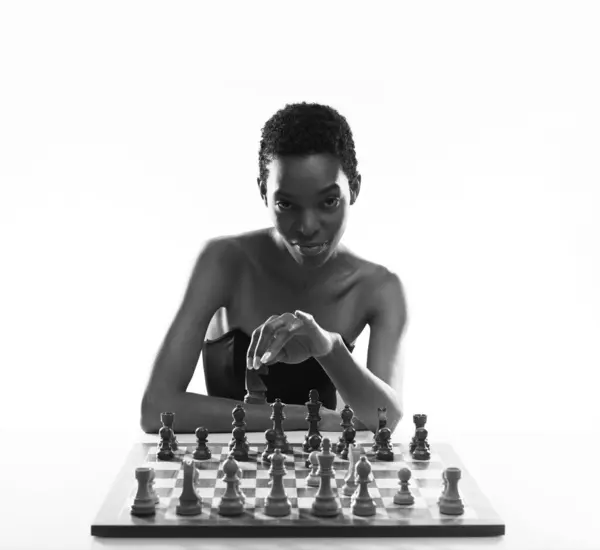 stock image Beautiful black woman playing chess, she is holding a piece and looking at camera, chess game concept