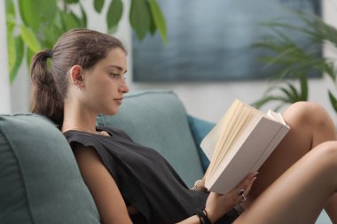 Güzel, rahat, genç bir kız evdeki kanepede dinleniyor ve kitap okuyor, yaşam tarzı konsepti.