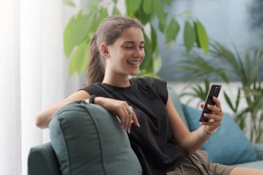 Şirin, mutlu bir genç, evdeki kanepede dinleniyor ve akıllı telefonuyla iletişim kuruyor, sosyal iletişim kuruyor ve sohbet ediyor, yaşam tarzı ve teknoloji kavramı.