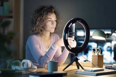 Masasında oturan ve takipçileri için bir video kaydetmeye hazırlanan genç bir kadın nüfuzlu kişi, akıllı telefonundaki ayarları kontrol ediyor.