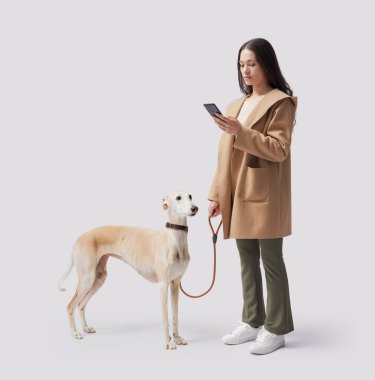 Genç bir kadın köpeğini gezdiriyor ve beyaz arka planda izole edilmiş akıllı telefonunu kontrol ediyor.