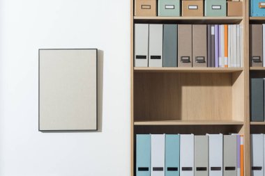 Kitaplıkta klasörler ve kitaplar olan düzenli ofis içi duvarda asılı boş sanat eserleri, depolama ve organizasyon konsepti