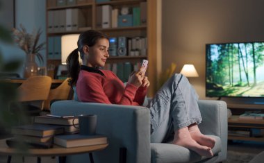 Oturma odasındaki koltukta oturan ve akıllı telefonunu kullanan güzel bir kız sosyal medyada sohbet ediyor ve oda içini kontrol ediyor.