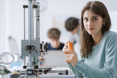 Laboratuvarda kendinden emin mutlu bir kız öğrencinin portresi. Üç boyutlu bir yazıcıyla çalışıyor ve kameraya gülümsüyor.