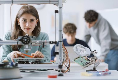 Laboratuvarda kendinden emin mutlu bir kız öğrencinin portresi. Üç boyutlu bir yazıcıyla çalışıyor ve kameraya gülümsüyor.