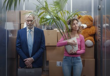 Woman moving into her new apartment, she is standing in the elevator surrounded by many cardboard boxes and holding a big houseplant, a man is standing next to her and feeling bothered clipart