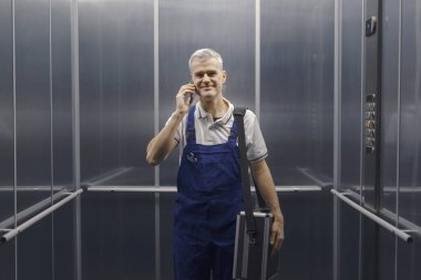 Gülümseyen teknisyen asansörde duruyor ve telefonla konuşuyor, profesyonel servis ve yardım kavramı.