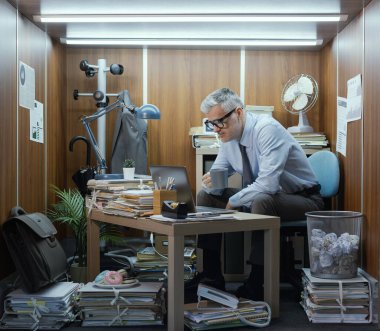 Sıkılmış ofis çalışanı küçük dağınık odasında kahve molası veriyor.