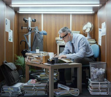 Verimli bir çalışan masasında oturuyor ve küçük bir ofiste çalışıyor. Kalemle yazıyor.