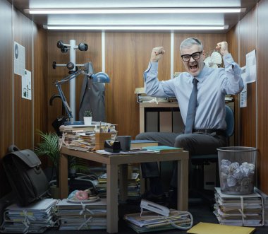 Küçük dağınık bir ofiste masasında oturan komik bir işadamının portresi, yumruklarını kaldırıp kameraya gülümsüyor.