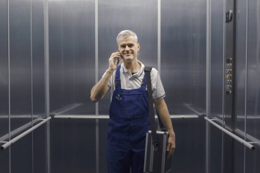 Gülümseyen teknisyen asansörde duruyor ve telefonla konuşuyor, profesyonel servis ve yardım kavramı.