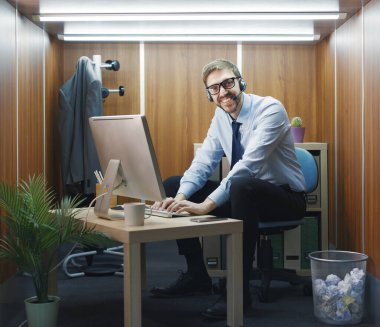 Masasında oturan ve küçük bir ofiste çalışan mutlu bir çalışan, bir kulaklık takıyor ve daktilo yazıyor.