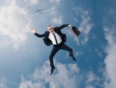 Arka planda bulutlar, gökyüzü ve uçaktan düşen evrak çantasıyla dehşete düşmüş bir iş adamı..