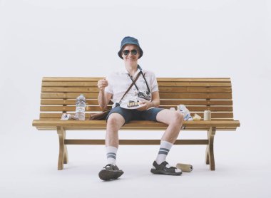 Beyaz arka planda çöplerle dolu bir bankta oturan kaba ve kaba turist..