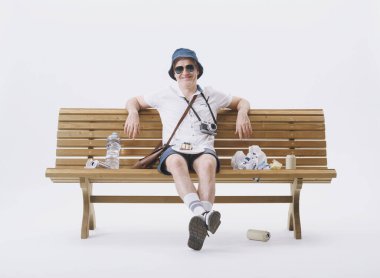 Beyaz arka planda çöplerle dolu bir bankta oturan kaba ve kaba turist..