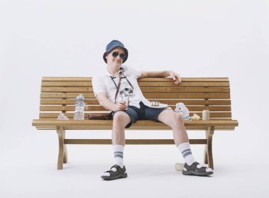 Beyaz arka planda çöplerle dolu bir bankta oturan kaba ve kaba turist..