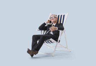 Siyah takım elbiseli orta yaşlı bir yönetici plaj sandalyesinde otururken telefonla konuşuyor. Tarafsız bir arkaplanda izole bir şekilde..