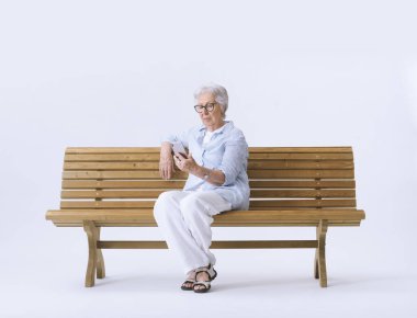 Yaşlı bir kadın bankta otururken telefonuna bakar..