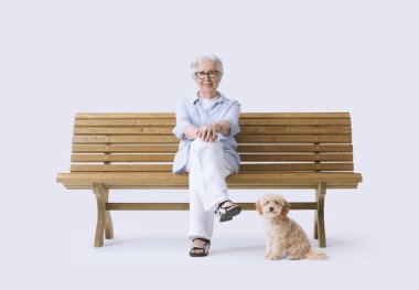 Gülümseyen ve mutlu yaşlı bir kadın köpeğiyle bankta oturuyor, kameraya bakıyor..