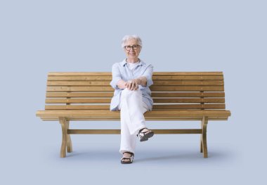 Gülümseyen ve mutlu yaşlı kadın bankta oturmuş kameraya bakıyor..