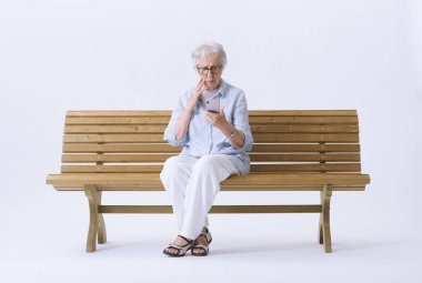 Yaşlı bir kadın bankta otururken endişeli bir şekilde telefonuna bakar. Kötü haber kavramı.