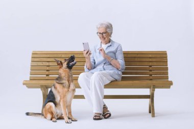 Gülümseyen ve mutlu yaşlı bir kadın köpeğiyle bankta oturuyor, telefonuna bakıyor..