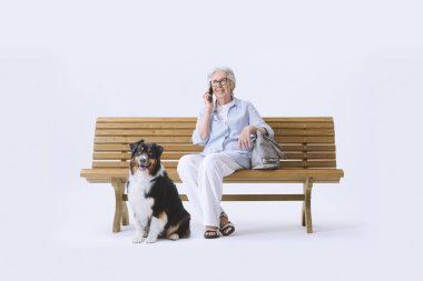 Gülümseyen ve mutlu yaşlı bir kadın köpeğiyle bankta oturuyor, telefonda konuşuyor..