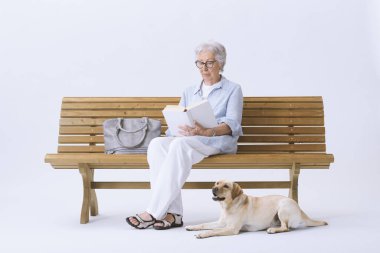 Yaşlı bir kadın köpeğiyle bankta oturmuş kitap okuyor..