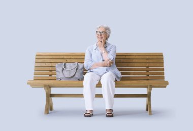 Gülümseyen ve mutlu yaşlı bir kadın bankta oturuyor, yukarı bakarken düşünüyor..