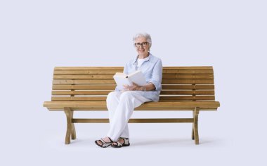 Yaşlı, gülümseyen ve mutlu bir kadın bankta oturmuş kitap okuyor..