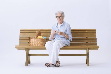 Gülümseyen ve mutlu yaşlı bir kadın bankta oturmuş örgü örüyor ve kameraya bakıyor..