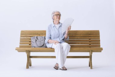 Yaşlı kadın bankta oturuyor, sıcaktan acı çekiyor ve kendini yelliyor. Küresel ısınma kavramı.