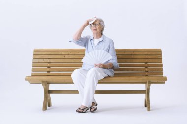 Yaşlı kadın bankta oturuyor, sıcaktan acı çekiyor ve kendini yelliyor. Küresel ısınma kavramı.