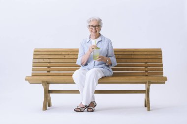 Gülümseyen ve mutlu yaşlı bir kadın bankta oturmuş elinde bir bardak su tutarak kameraya bakıyor..