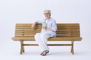 Gülümseyen ve mutlu yaşlı bir kadın bankta oturmuş elinde bir bardak su tutarak kameraya bakıyor..