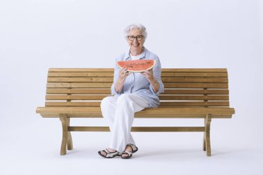 Yaşlı, gülümseyen ve mutlu bir kadın bankta oturmuş kameraya bakarak bir dilim karpuz yiyor..