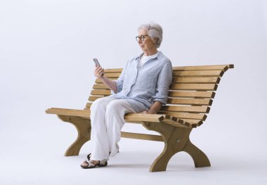 Yaşlı bir kadın bankta otururken telefonuna bakar..