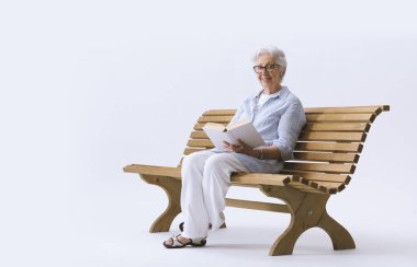 Yaşlı, gülümseyen ve mutlu bir kadın bankta oturmuş kitap okuyor..