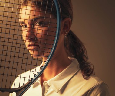 Beyaz elbiseli esmer tenisçinin yakın plan portresi. Tarafsız bir arkaplana karşı poz vermiş. Güzelliği, zarafeti ve atletik stili vurguluyor..