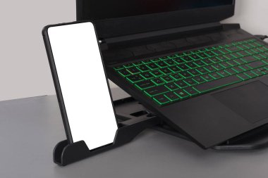 Modern gaming laptop with green backlit keyboard and a smartphone with blank screen on a holder clipart