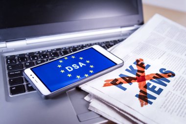 DSA ve AB bayrağını taşıyan akıllı telefon sahte haber ve dizüstü bilgisayarlı bir gazete ekranında. Avrupa Birliği Dijital Hizmet Yasası veya DSA, gerçeklerin kontrol edilmesi de dahil olmak üzere proaktif ılımlılığı düzenler