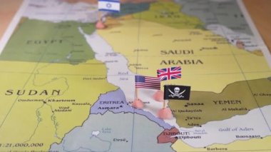 ABD ve İran 'ın bayrakları Kızıl Deniz bölgesinin haritasında bir korsan amblemini çevreleyen bayraklar. Bu stratejik çatışmalardaki karmaşık jeopolitik dinamikleri ve potansiyel çatışmaları sembolik olarak temsil eder.