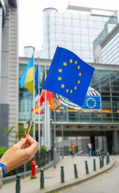 Belçika 'nın Brüksel kentindeki Avrupa Parlamentosu binasının önünde küçük bir Avrupa Birliği bayrağını tutan bir el resmi. Arka planda başka bayraklar ve modern mimari