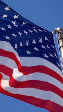 Açık mavi gökyüzüne karşı rüzgarda dalgalanan Amerikan bayrağının canlı yakın çekimi. Bu resim, vatanseverliği simgeleyen ABD bayrağının ikonik kırmızı, beyaz ve mavi renklerini yakalar.